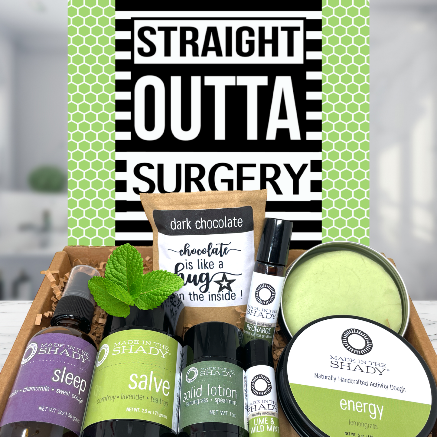 A curated gift box set from Shady Apothecary featuring multiple scented products including sleep spray, herbal salve, solid lotion, energy balm, and a dark chocolate candle, arranged in a gift box in front of a green and black patterned card that reads 'STRAIGHT OUTTA SURGERY'.