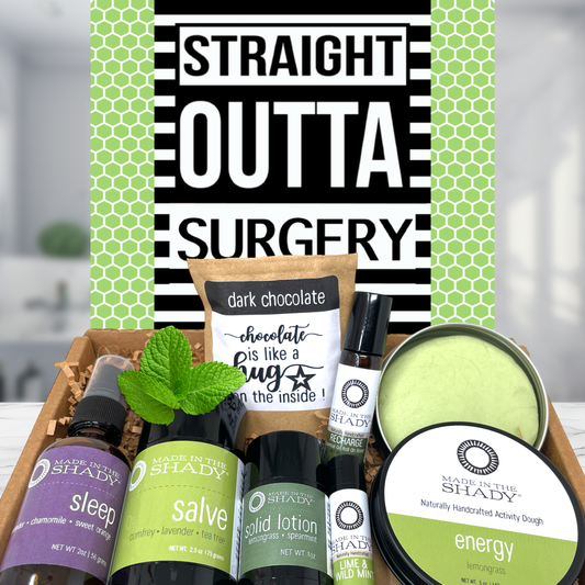 A curated gift box set from Shady Apothecary featuring multiple scented products including sleep spray, herbal salve, solid lotion, energy balm, and a dark chocolate candle, arranged in a gift box in front of a green and black patterned card that reads 'STRAIGHT OUTTA SURGERY'.